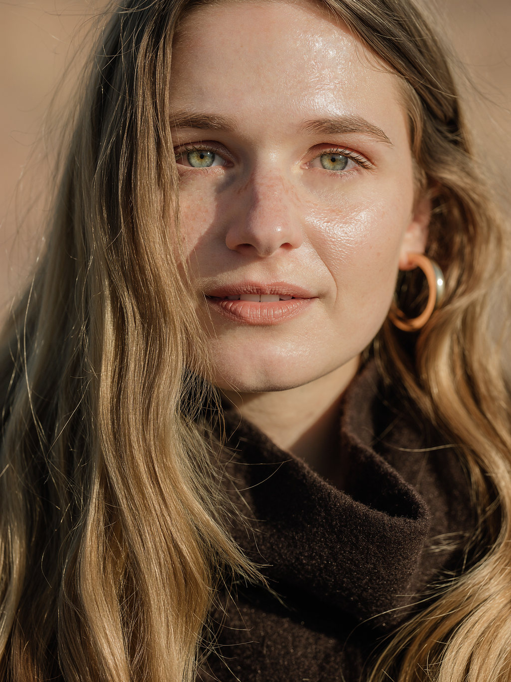 Close portrait of Lilyanna in a chocolate turtleneck and sculptural gold hoop earrings, lit by warm late sun.