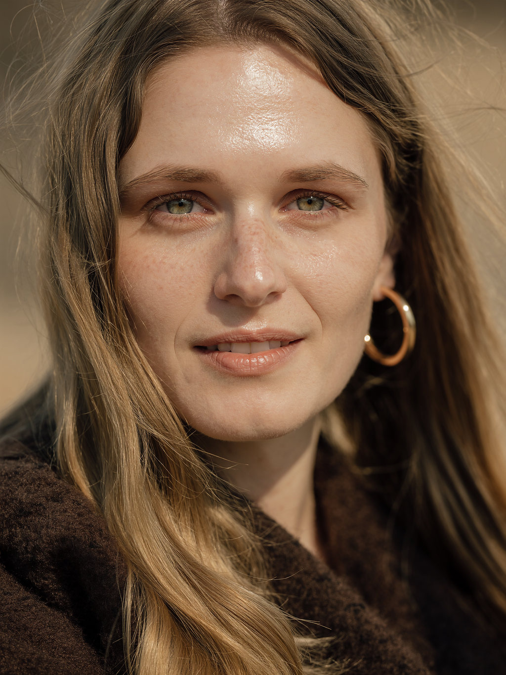 Lilyanna smiling softly in a brown fleece collar and polished rose-gold hoops, warm light on freckled skin.