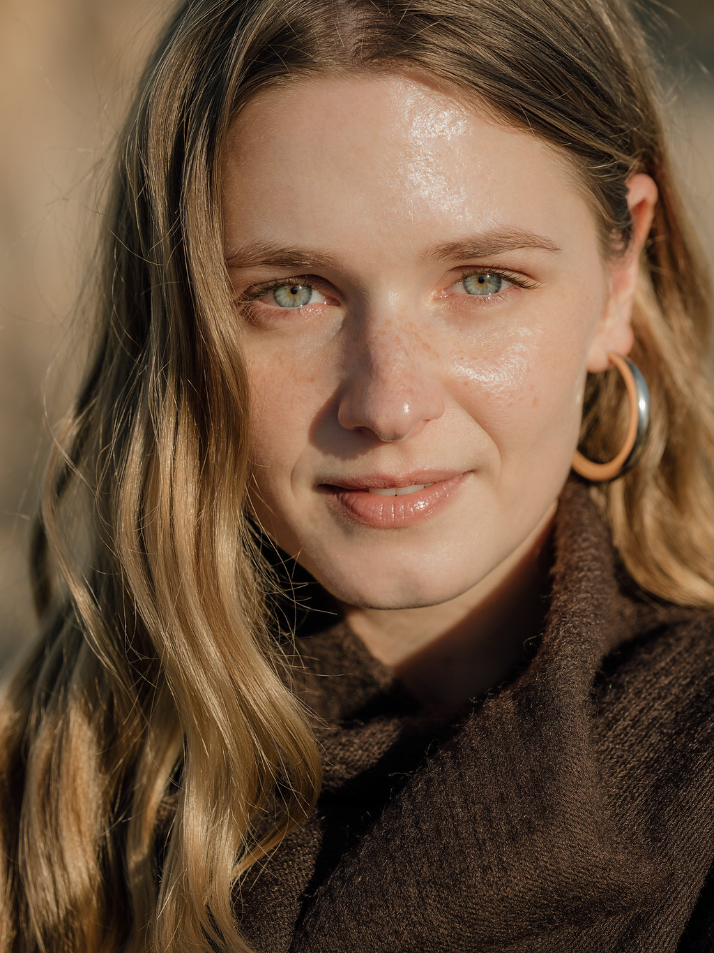 Lilyanna smiling in a brown turtleneck with silver and gold hoop earrings, lit by warm sun.