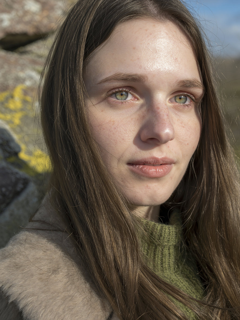 Lilyanna gazing into the distance beside lichen-covered stone, sunlight on freckled skin.