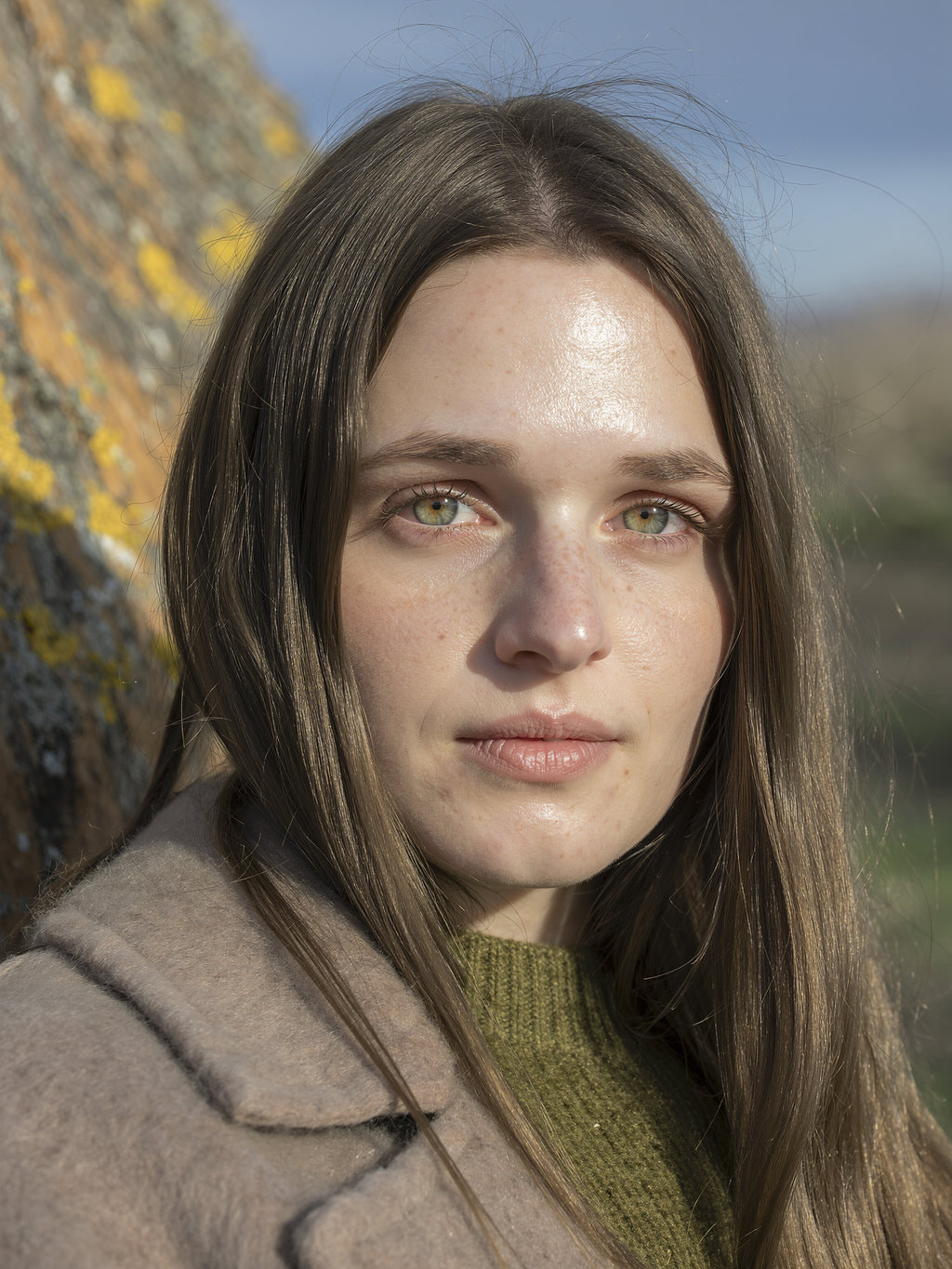 Portrait of Lilyanna against a lichen-covered rock in a taupe wool coat and olive knit.