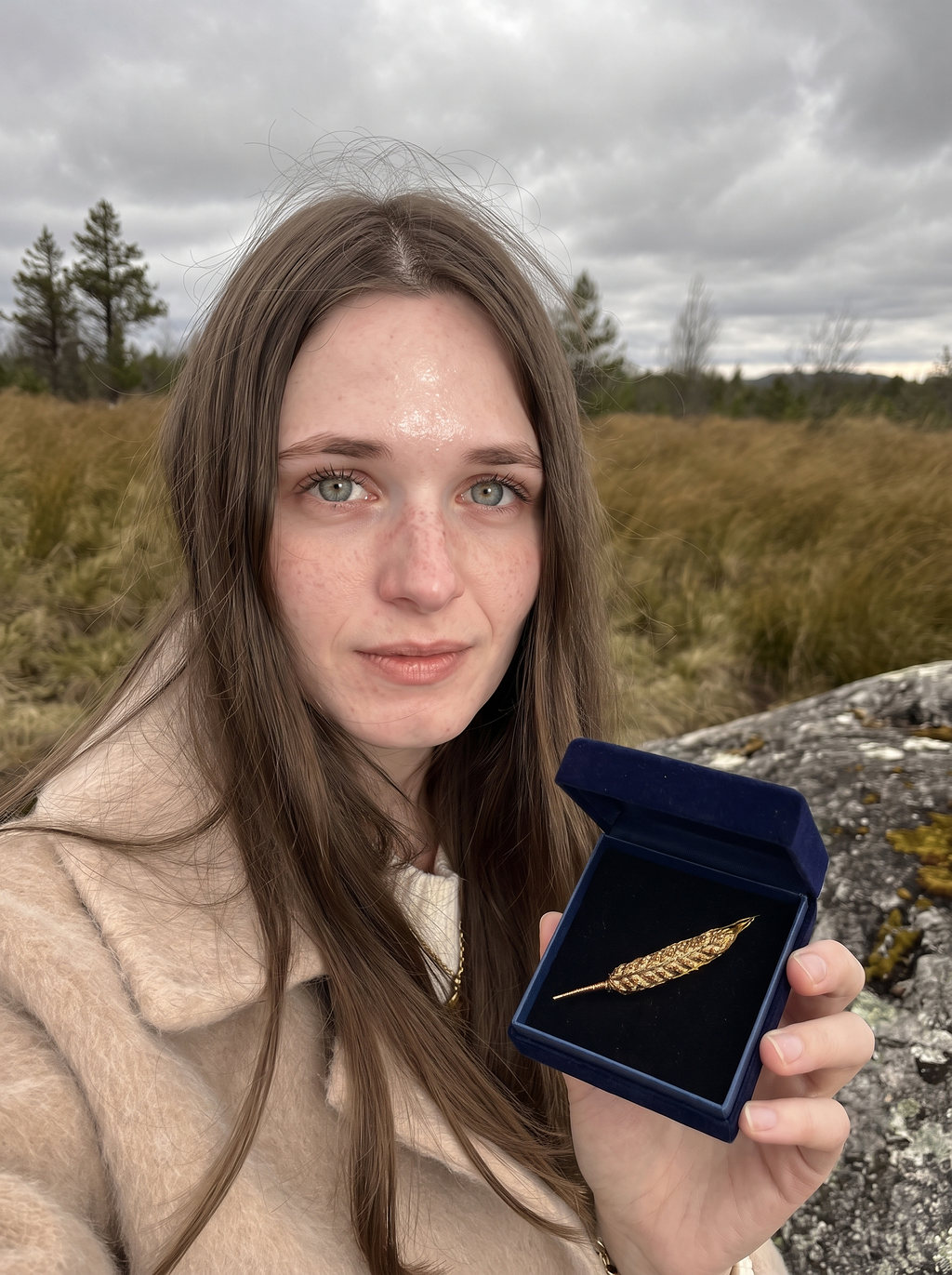 Lilyanna on a windswept moor holding an open navy velvet box with a gold feather brooch.