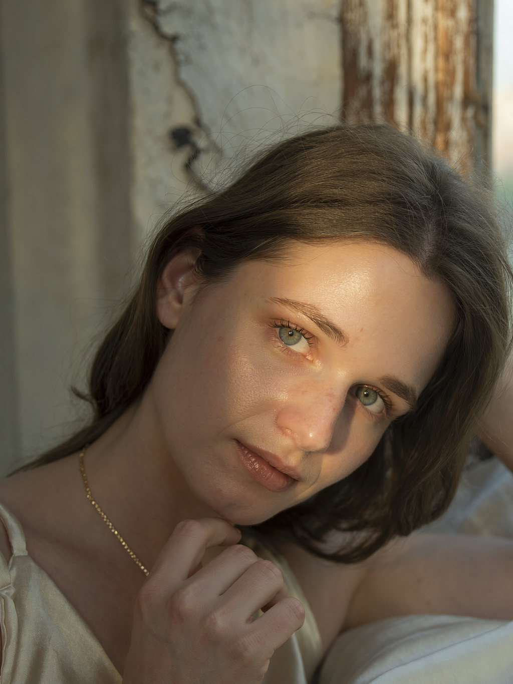 Lilyanna resting her chin on her hand by a weathered window, a fine gold chain and satin camisole in low sun.